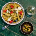 a white bowl filled with thick rigatoni pasta, green pesto, red cherry tomatoes, creamy burrata cheese, caper berries and a glass of white wine sits on top of a green wooden dining table
